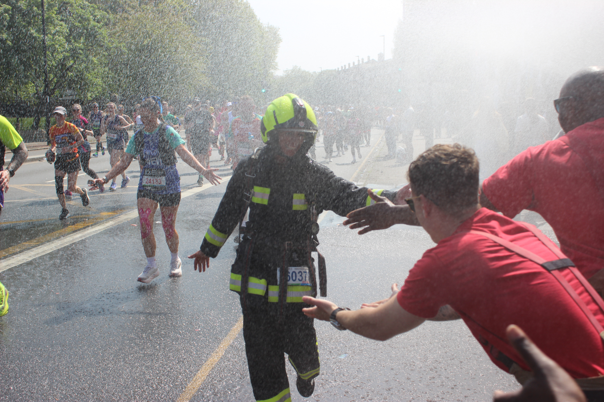 A firefighter in gear runs through mist and high-fives a person in a red shirt during a marathon, with other runners and sprays of water in the background on a sunny day.
