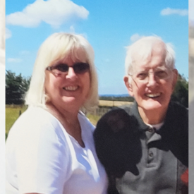 A smiling woman with blonde hair and sunglasses stands next to an elderly man with white hair and glasses outdoors on a sunny day, with a blue sky and green trees in the background.