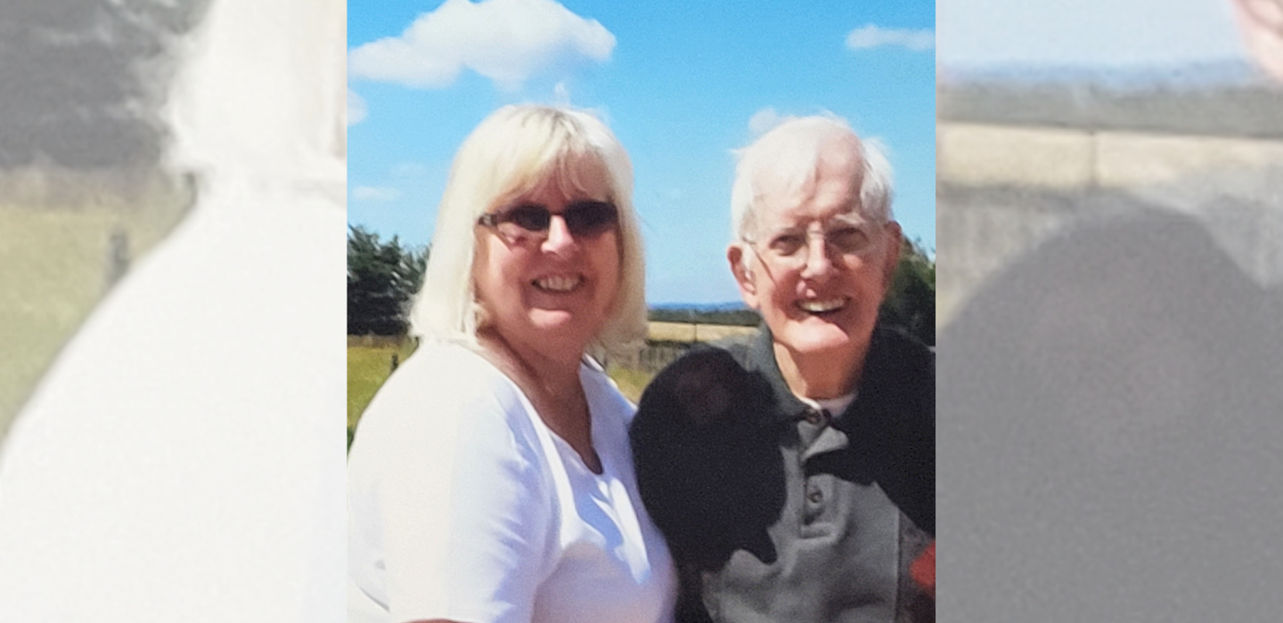 A smiling woman with blonde hair and sunglasses stands next to an elderly man with white hair and glasses outdoors on a sunny day, with a blue sky and green trees in the background.