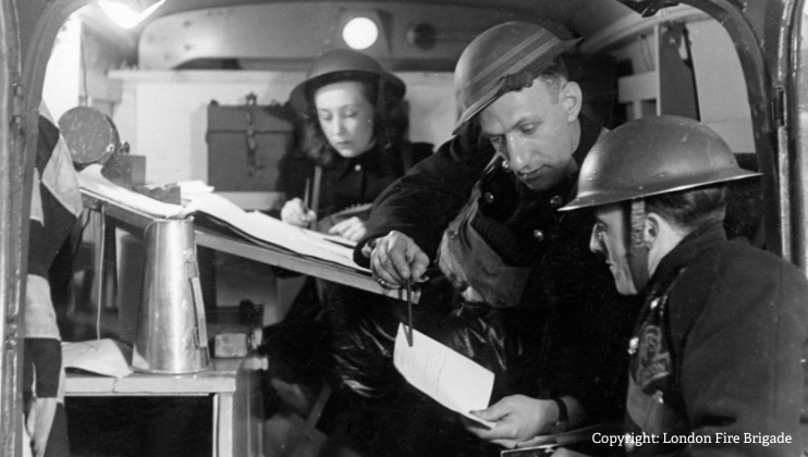 Three people wearing World War II-era helmets are inside a command vehicle, examining papers and writing notes, focused on their work. A woman is seated in the background, and two men are in the foreground.