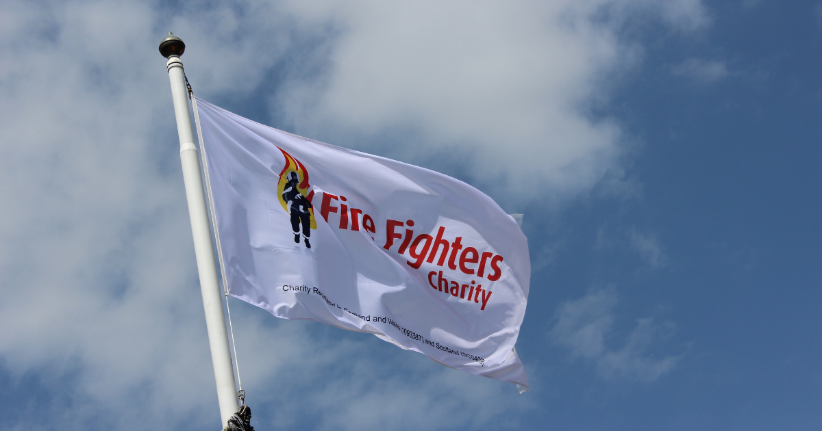 A white flag with the Fire Fighters Charity logo featuring a firefighter and flames, flying on a pole against a partly cloudy blue sky.