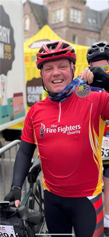 A smiling cyclist wearing a red "The Fire Fighters Charity" jersey and helmet holds up a medal, standing beside a bicycle at an outdoor event with banners and buildings in the background.
