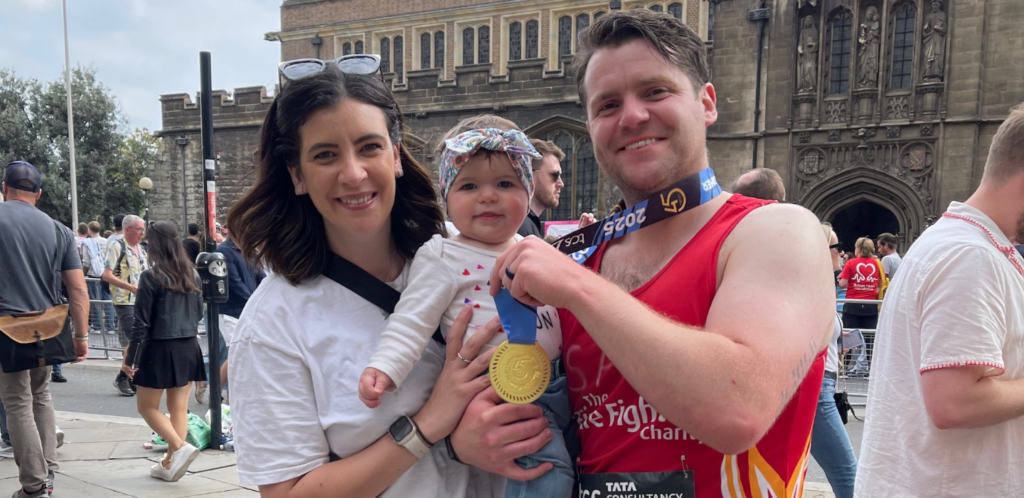 A smiling woman and man stand outdoors with a baby, who wears a headband. The man proudly holds up a race medal, wearing athletic gear. People and an old stone building are in the background.