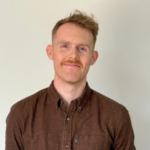 A man with short red hair and a mustache, wearing a buttoned-up brown shirt, stands against a plain off-white background. The image has a red border.