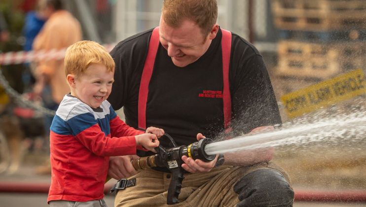 A smiling firefighter kneels and helps a young boy spray water from a fire hose. The boy looks excited as he grips the hose, with mist spraying out, and the firefighter supports him, both enjoying the moment outdoors.