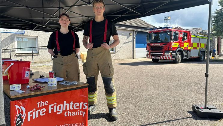 Two firefighters in uniform stand under a canopy behind a table for The Fire Fighters Charity. A fire engine is parked nearby, and donation items are displayed on the table. Buildings are visible in the background.
