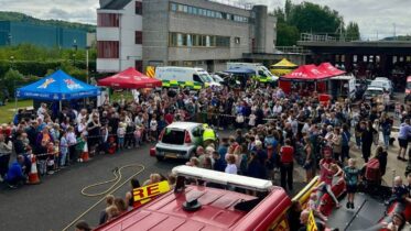A large crowd gathers at an outdoor emergency services event featuring tents, emergency vehicles, and a demonstration with a car. People watch from behind barriers on a cloudy day near a large building.