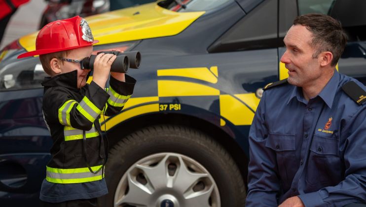 A young child in a firefighter costume looks through binoculars, while an adult in a blue uniform kneels nearby and smiles at the child, with a yellow and blue emergency vehicle in the background.