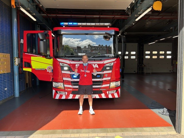 A smiling man in a red shirt stands in front of a red fire engine inside a fire station garage, holding two blue trophies and wearing medals around his neck. The fire engine has "Scottish Fire" written on its door.