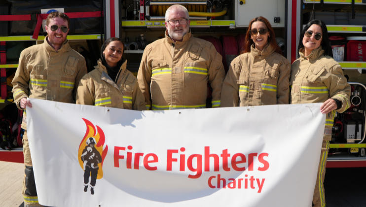 Five firefighters in uniform stand in front of a fire truck, smiling and holding a banner that reads "Fire Fighters Charity" with a firefighter logo on it.