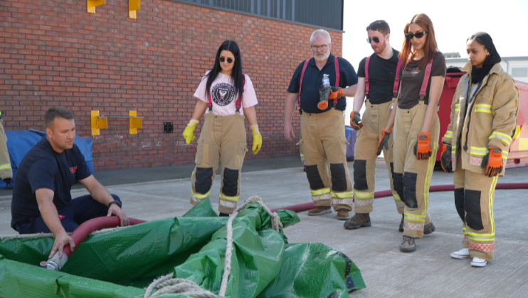 A group of five people, some in firefighting gear, watch as a firefighter kneels and handles a red hose near a green tarp on a concrete surface outside a brick building.
