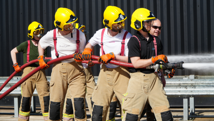 Four firefighters in yellow helmets and beige gear spray water from a fire hose, working together in a line outdoors in front of a black corrugated metal wall.