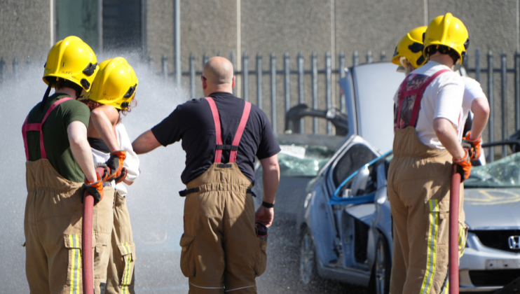 Four firefighters in yellow helmets and tan uniforms spray water near a damaged car, while a bald instructor in similar attire gives directions. A metal fence and building are visible in the background.