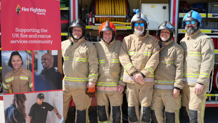 Six firefighters in uniform and helmets stand smiling in front of a fire truck. To the left is a banner for the Fire Fighters Charity, supporting the UK fire and rescue services community, with photos of staff and beneficiaries.