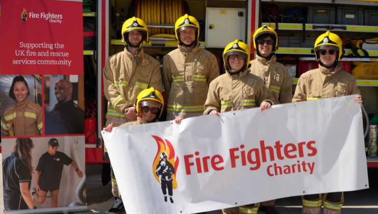 Six firefighters in full gear stand in front of a fire truck holding a “Fire Fighters Charity” banner, with a charity sign beside them supporting the UK fire and rescue services community.