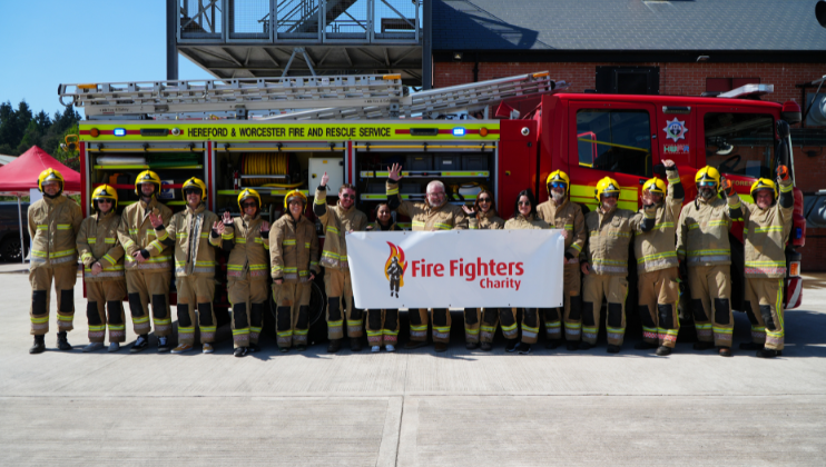 A group of firefighters in uniform stand in front of a fire engine, smiling and holding a large banner that reads