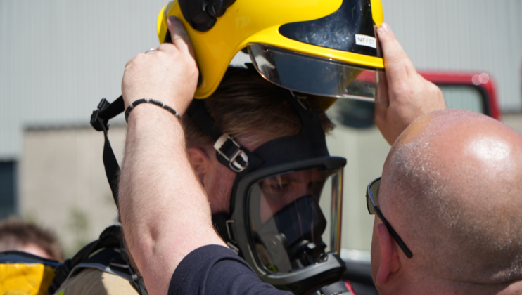 A person wearing a breathing mask is being assisted by another individual who is placing a yellow firefighter helmet on their head, likely as part of safety gear preparation.