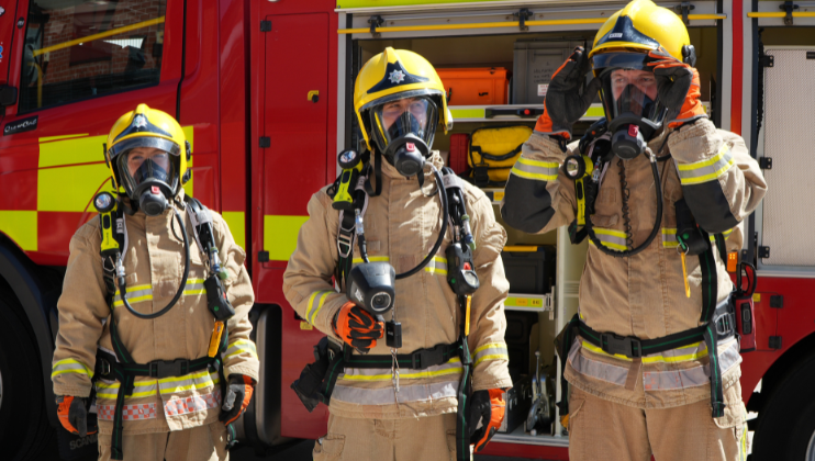 Three firefighters in full protective gear and helmets stand in front of a fire truck with open compartments, ready for action. Two face forward, and one adjusts their helmet.