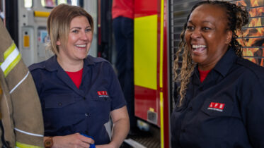 Two women firefighters wearing navy uniforms and smiling stand beside a fire truck. One holds a blue item, and both appear happy and engaged in conversation.