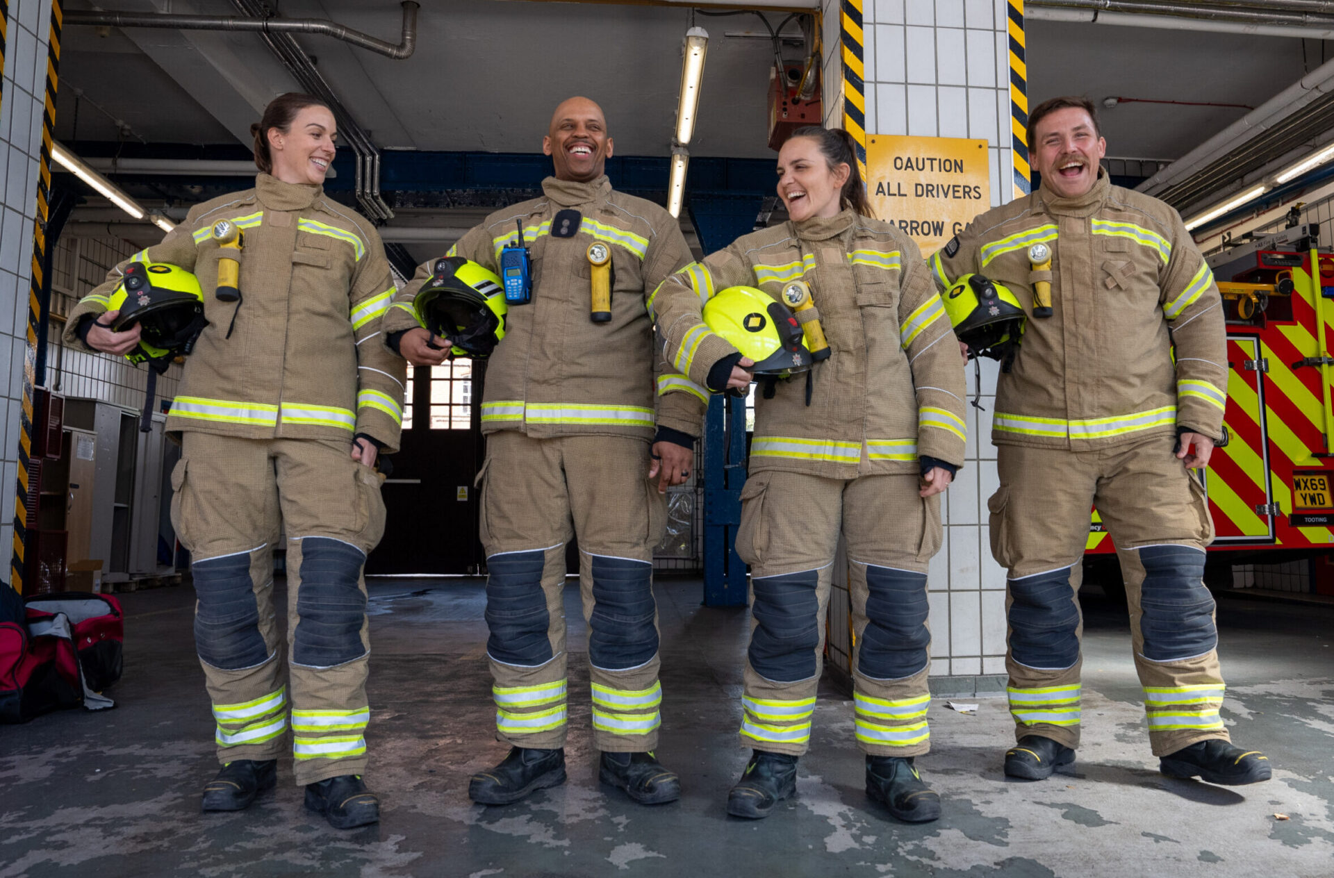 Four firefighters in uniform stand together smiling and holding helmets in a fire station garage, with a fire truck and caution signs in the background.