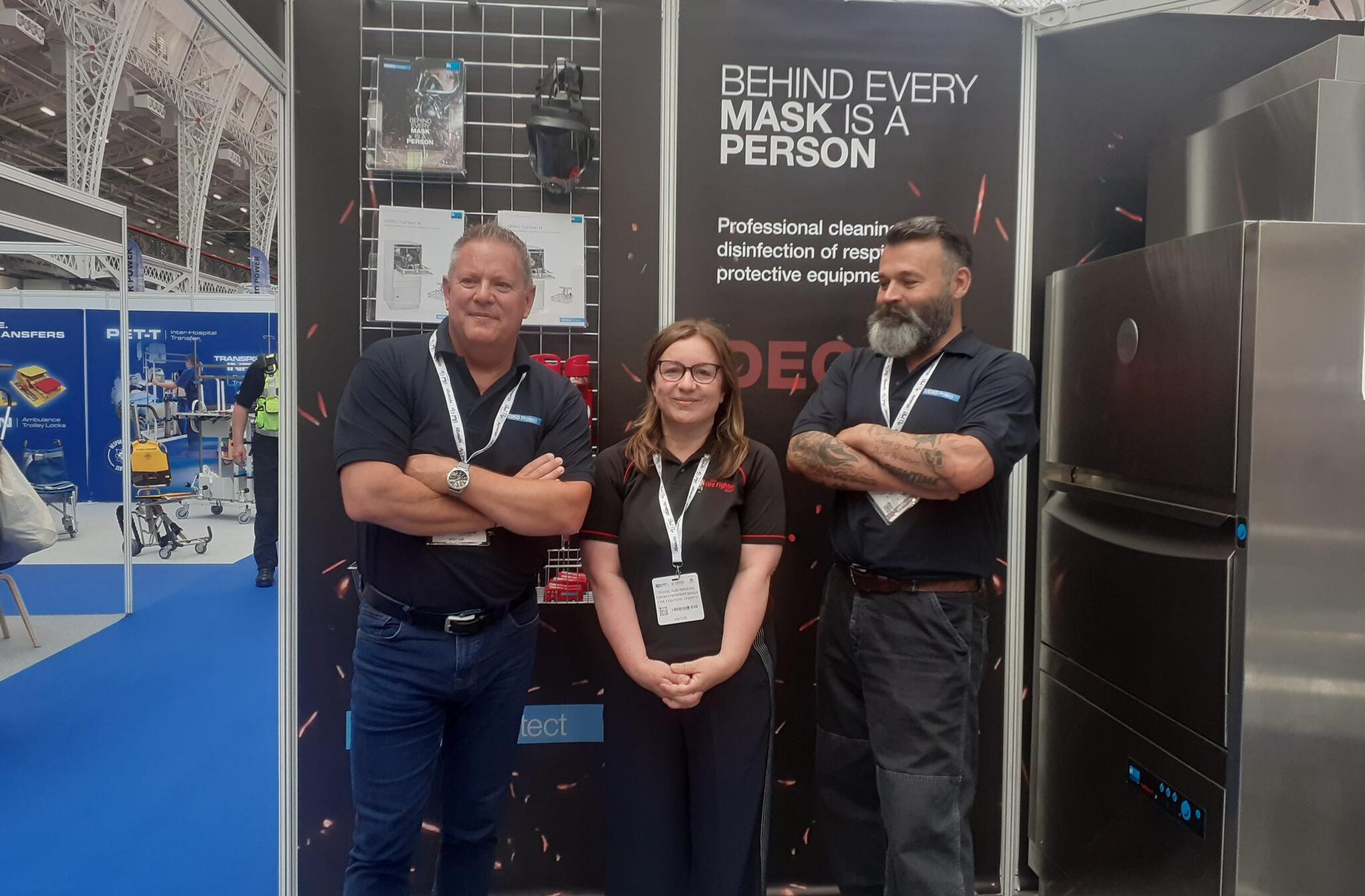 Three people wearing dark polo shirts and event lanyards stand in front of an exhibition booth displaying cleaning and disinfection equipment. A sign behind them reads,