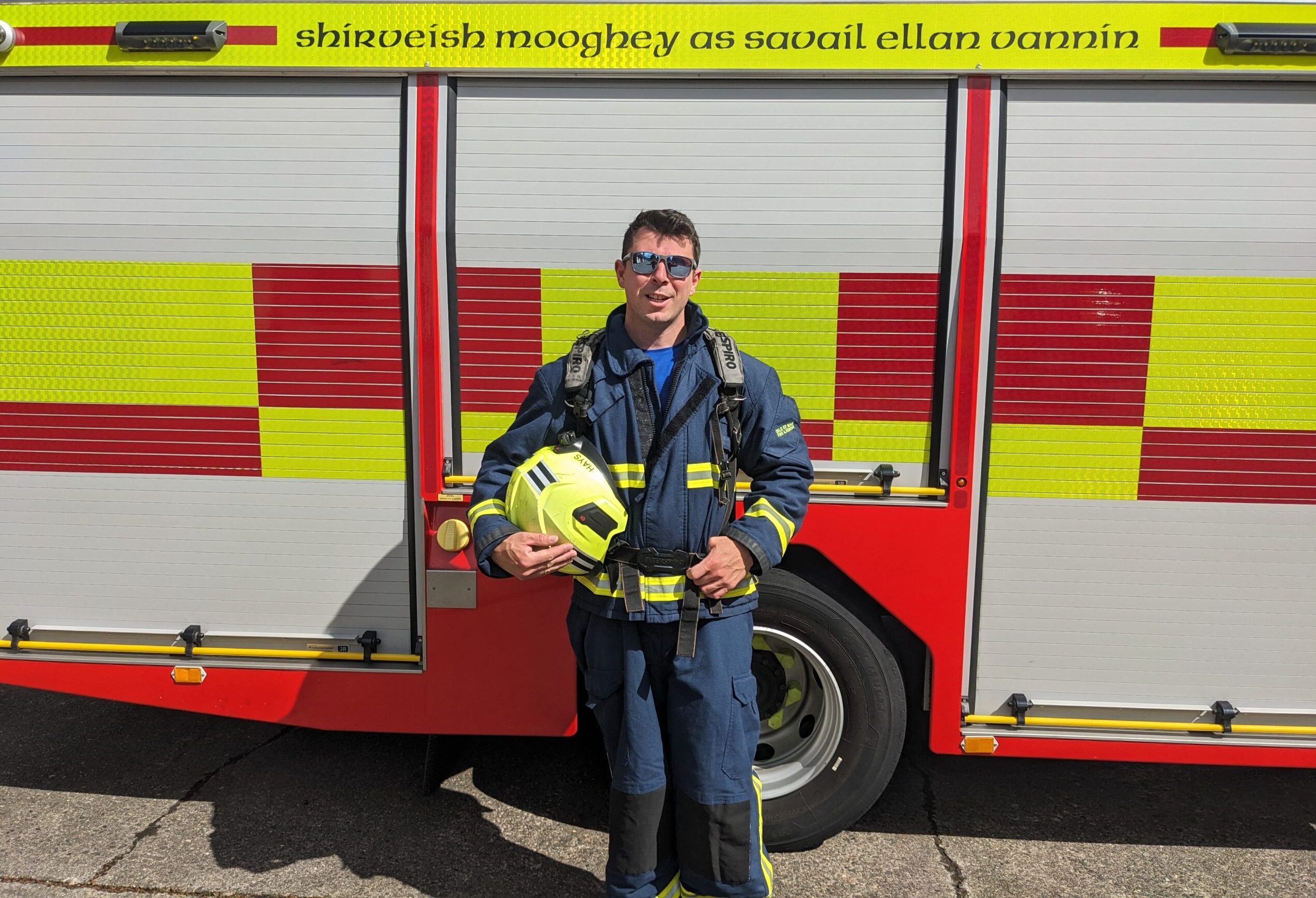 A firefighter in full uniform stands in front of a fire truck, holding a yellow helmet and smiling at the camera. The truck has red and yellow checkered patterns and text in a foreign language above the doors.