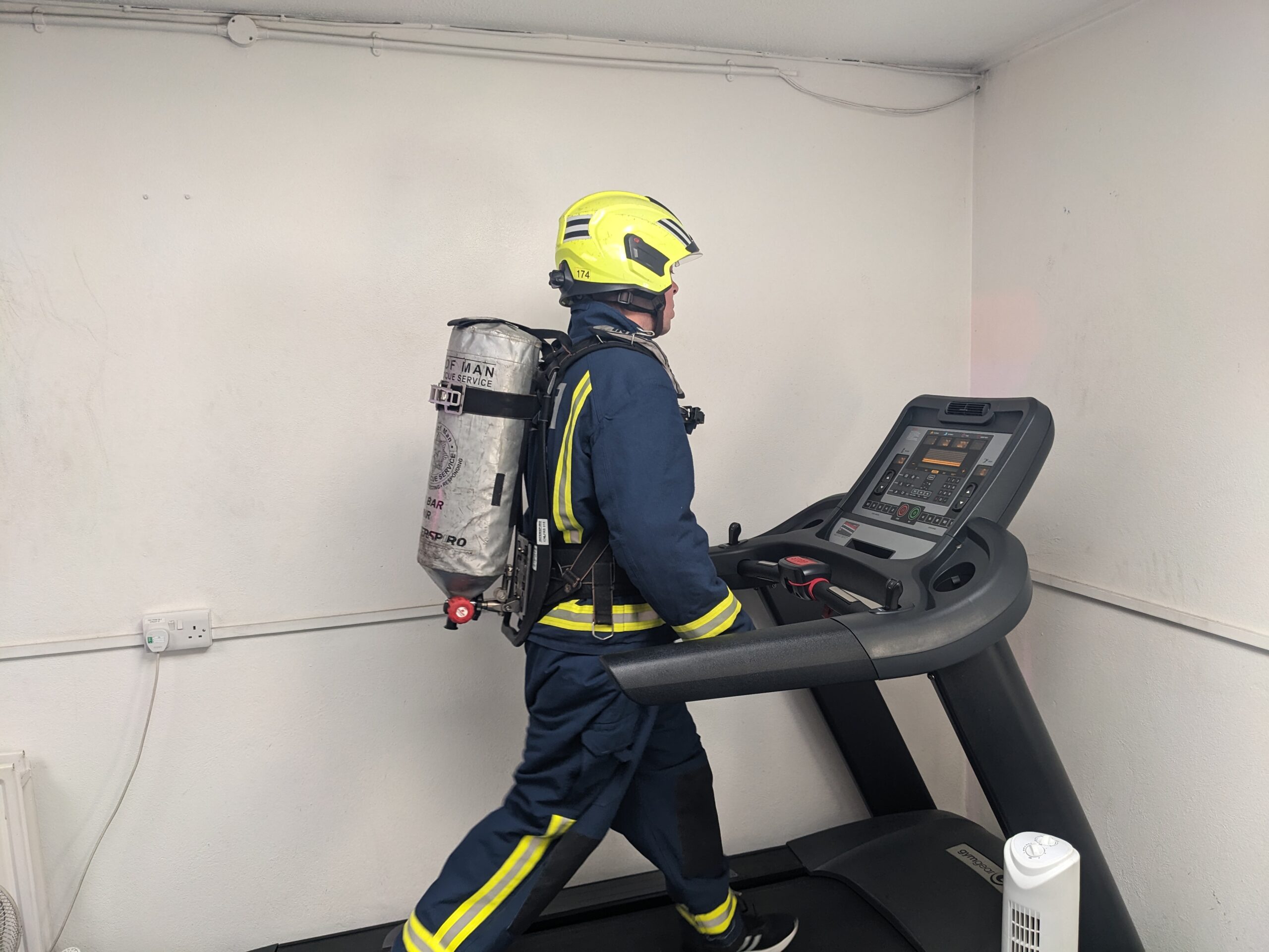 A firefighter in full protective gear and helmet walks on a treadmill indoors, wearing a large oxygen tank on their back.