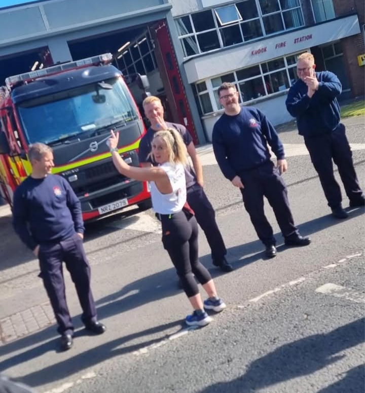 A woman gestures energetically while standing in front of five smiling firefighters outside a fire station with a fire truck parked nearby. The weather is sunny and bright.