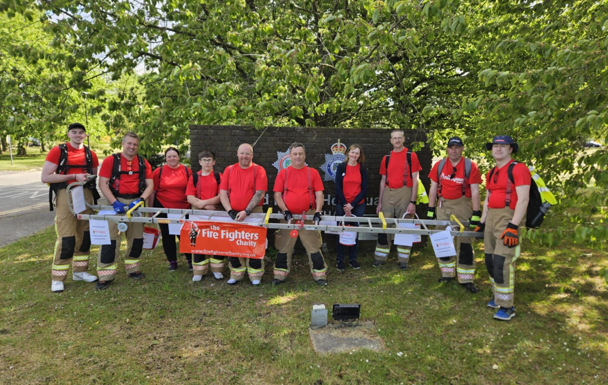 A group of firefighters in red shirts and gear stand outside in front of a leafy tree and a sign, holding a banner that reads