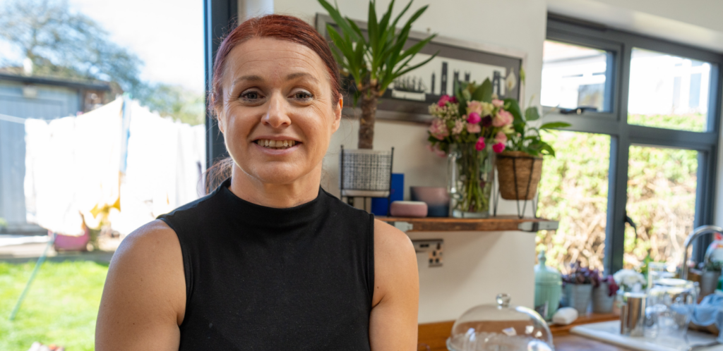 A woman with red hair in a ponytail, wearing a sleeveless black top, smiles in a bright kitchen with plants, flowers, and outdoor light streaming in through large windows.