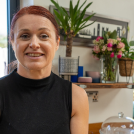 A woman with red hair in a ponytail, wearing a sleeveless black top, smiles in a bright kitchen with plants, flowers, and outdoor light streaming in through large windows.