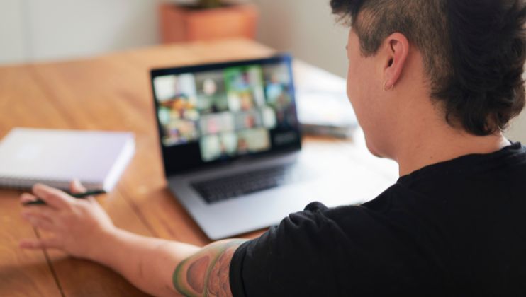 Person with short dark hair and a tattooed arm sits at a wooden table, attending a video call on a laptop. A closed notebook is nearby. The computer screen shows a grid of blurred people in a virtual meeting.