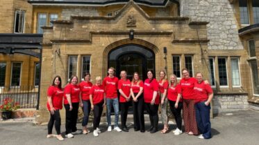 A group of twelve people stand outside a stone building, all wearing matching red shirts and smiling at the camera. Some people have their arms around each other, and the weather appears sunny.
