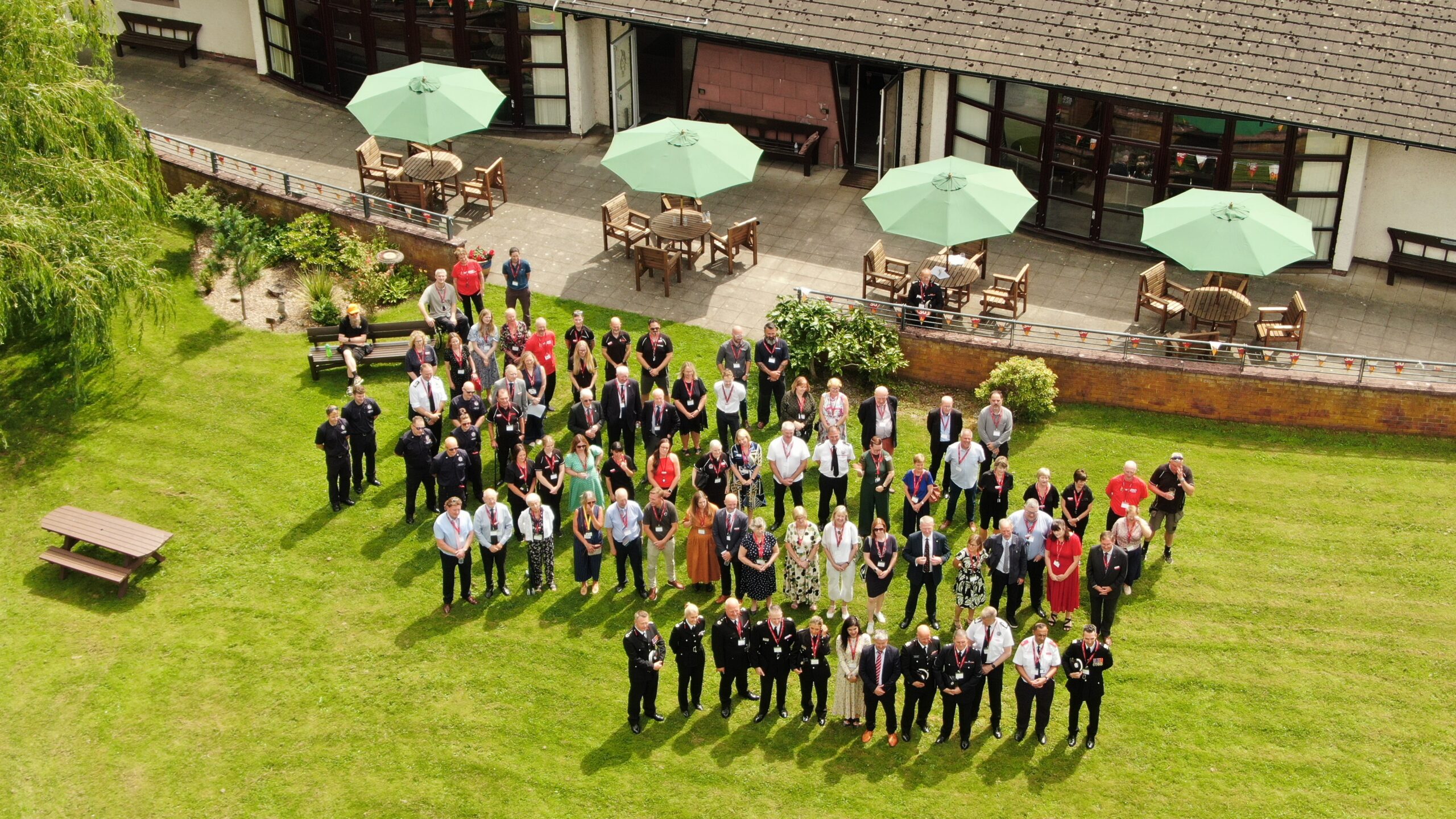 A large group of people stands together on a green lawn outside a building with outdoor seating and green umbrellas, posing for a photo on a sunny day.