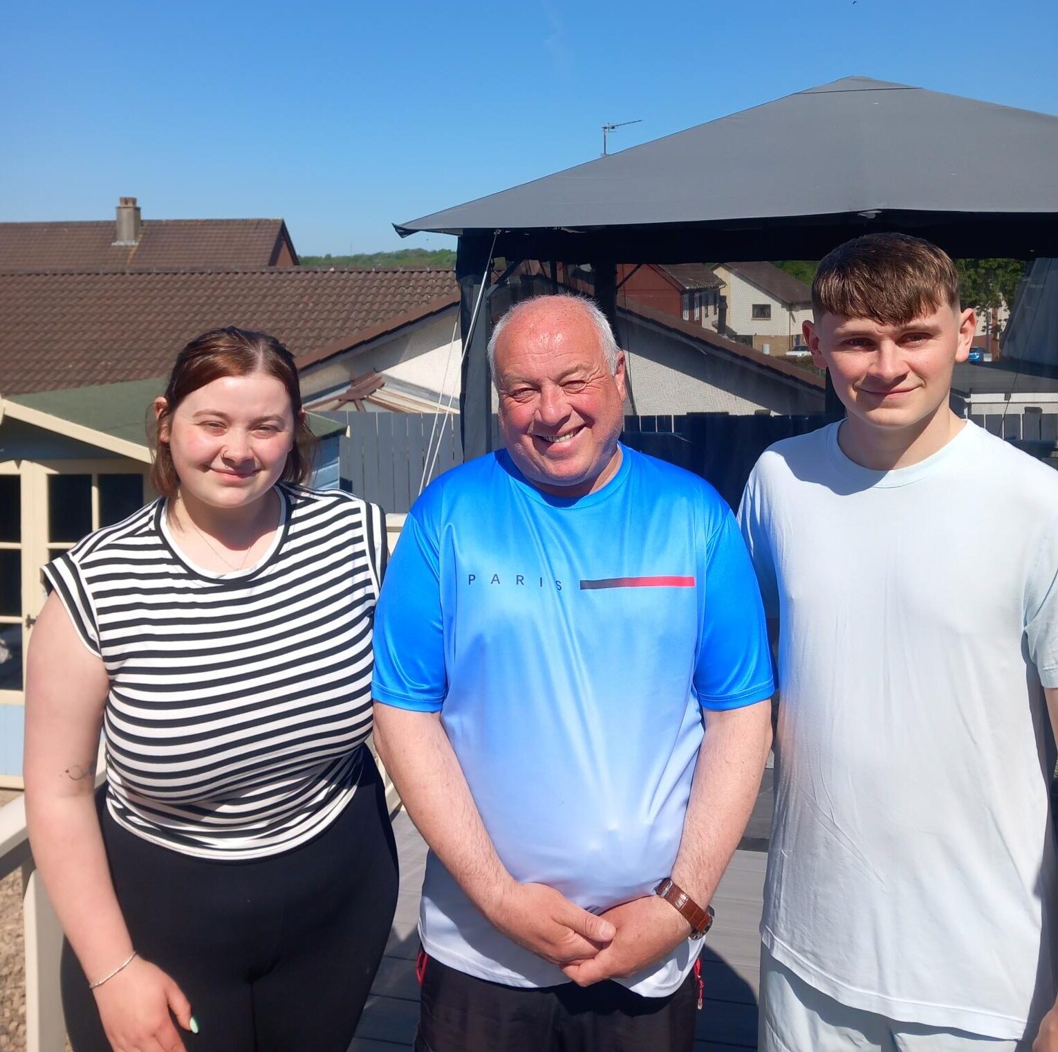 Three people stand smiling outdoors on a sunny day. A young woman in a black and white striped shirt is on the left, an older man in a blue shirt is in the center, and a young man in a light shirt is on the right. Houses and a gazebo are in the background.