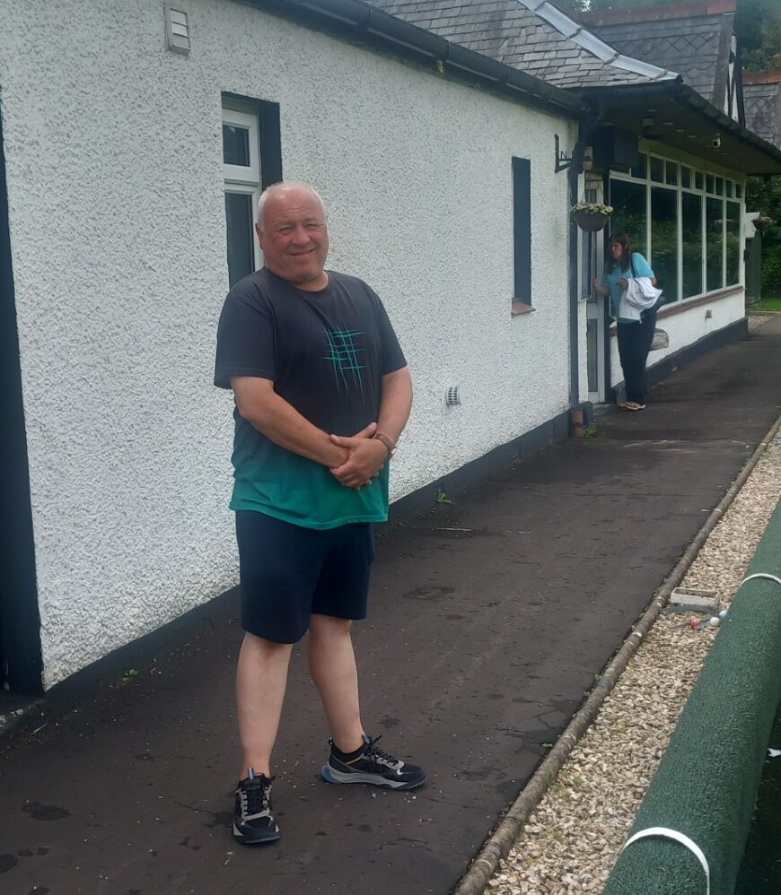 A man stands on a path beside a white building next to a green lawn, with another person visible near the building entrance in the background. The sky is overcast.
