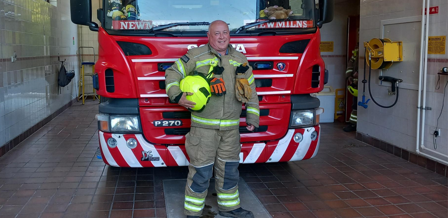 A smiling firefighter in full gear stands holding a yellow helmet in front of a red fire truck inside a fire station. The truck has white stripes and equipment visible in the background.