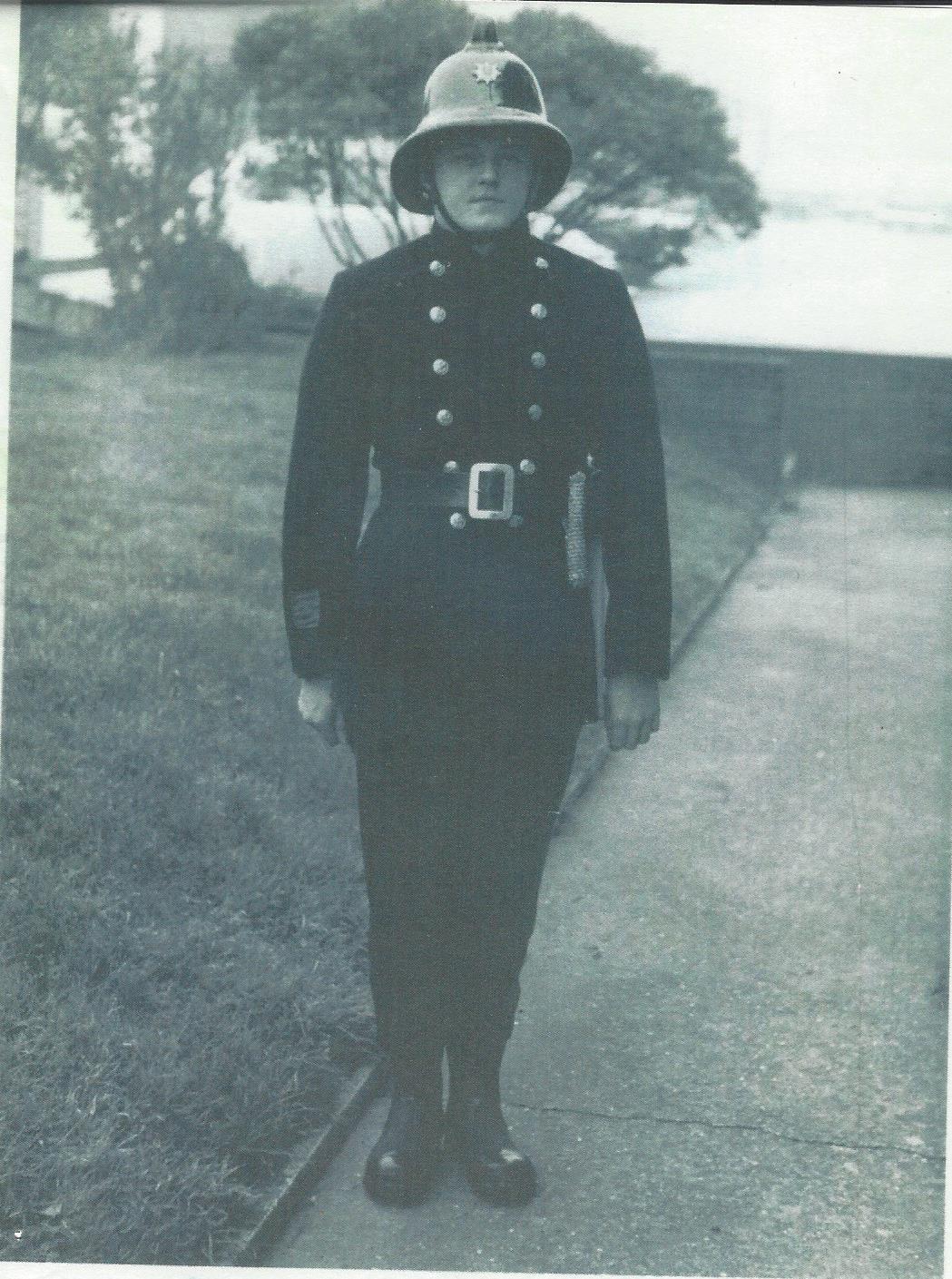 A person in an old-fashioned police uniform and helmet stands at attention on a paved path, with grass and trees in the background. The image is in black and white.