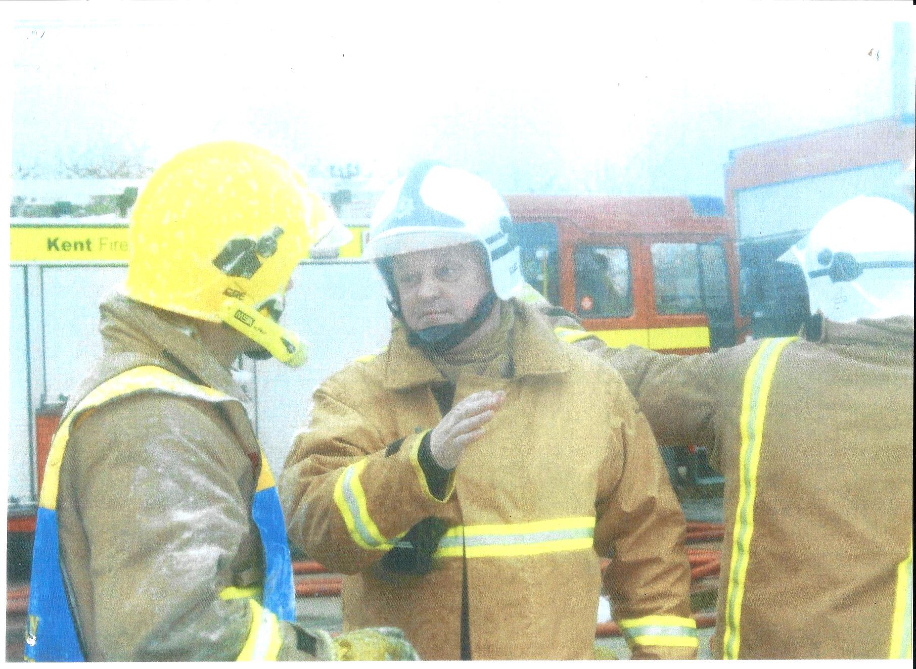 Three firefighters in protective gear and helmets stand and talk near fire trucks, with one firefighter raising a hand as if giving instructions. The scene appears to be at an emergency response site.