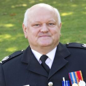 A man in a formal uniform with medals sits on a bench outdoors, holding a framed certificate and smiling at the camera. Green grass is visible in the background.