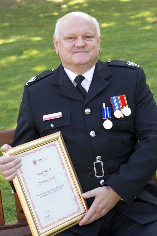 A man in a formal black uniform with medals sits on a bench outdoors, holding a framed certificate and smiling at the camera. Grass and sunlight are visible in the background.