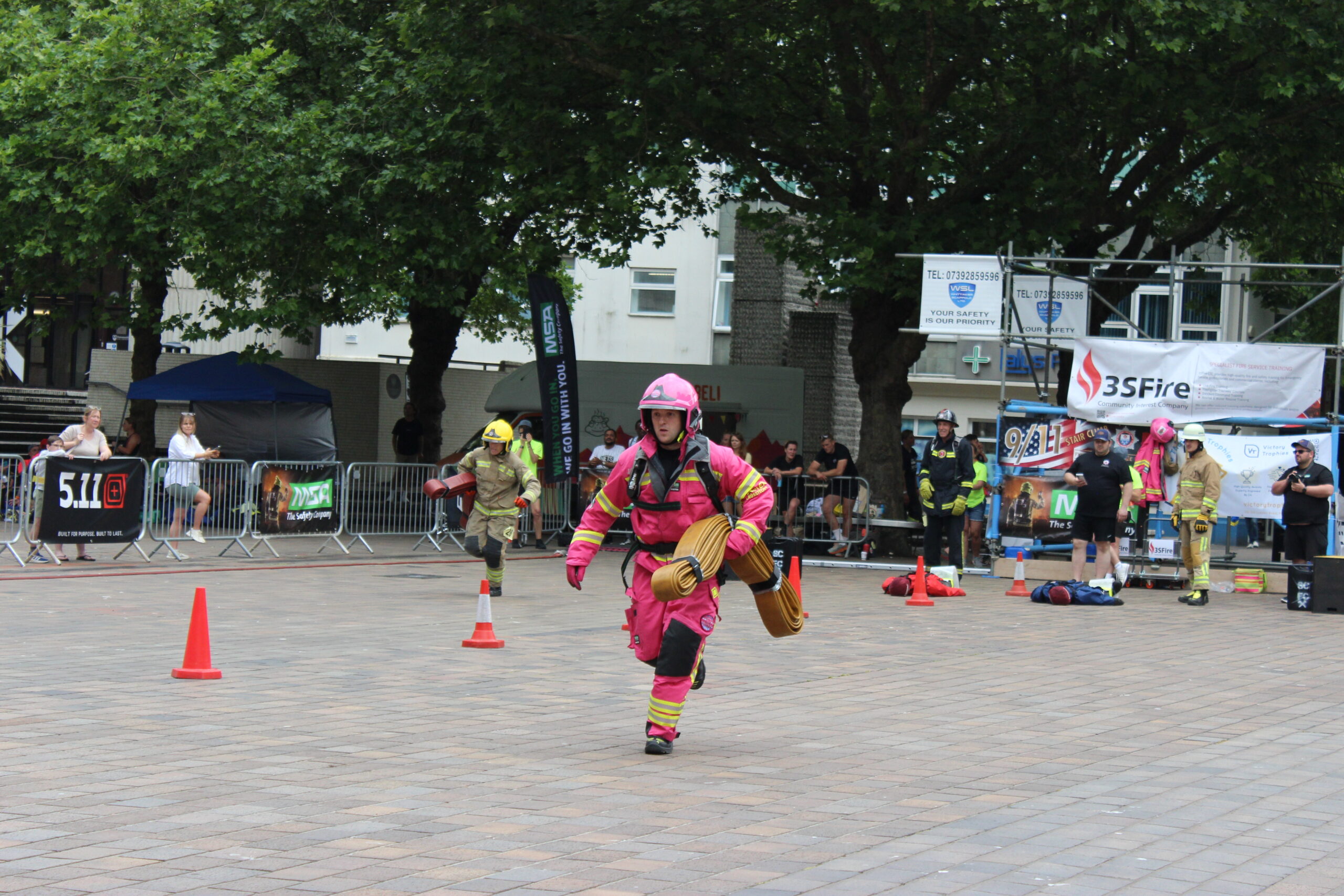 A firefighter in a bright pink uniform and helmet runs across a paved courtyard carrying a coiled hose, while other firefighters in standard gear participate in activities nearby. Orange cones and spectators are visible in the background.