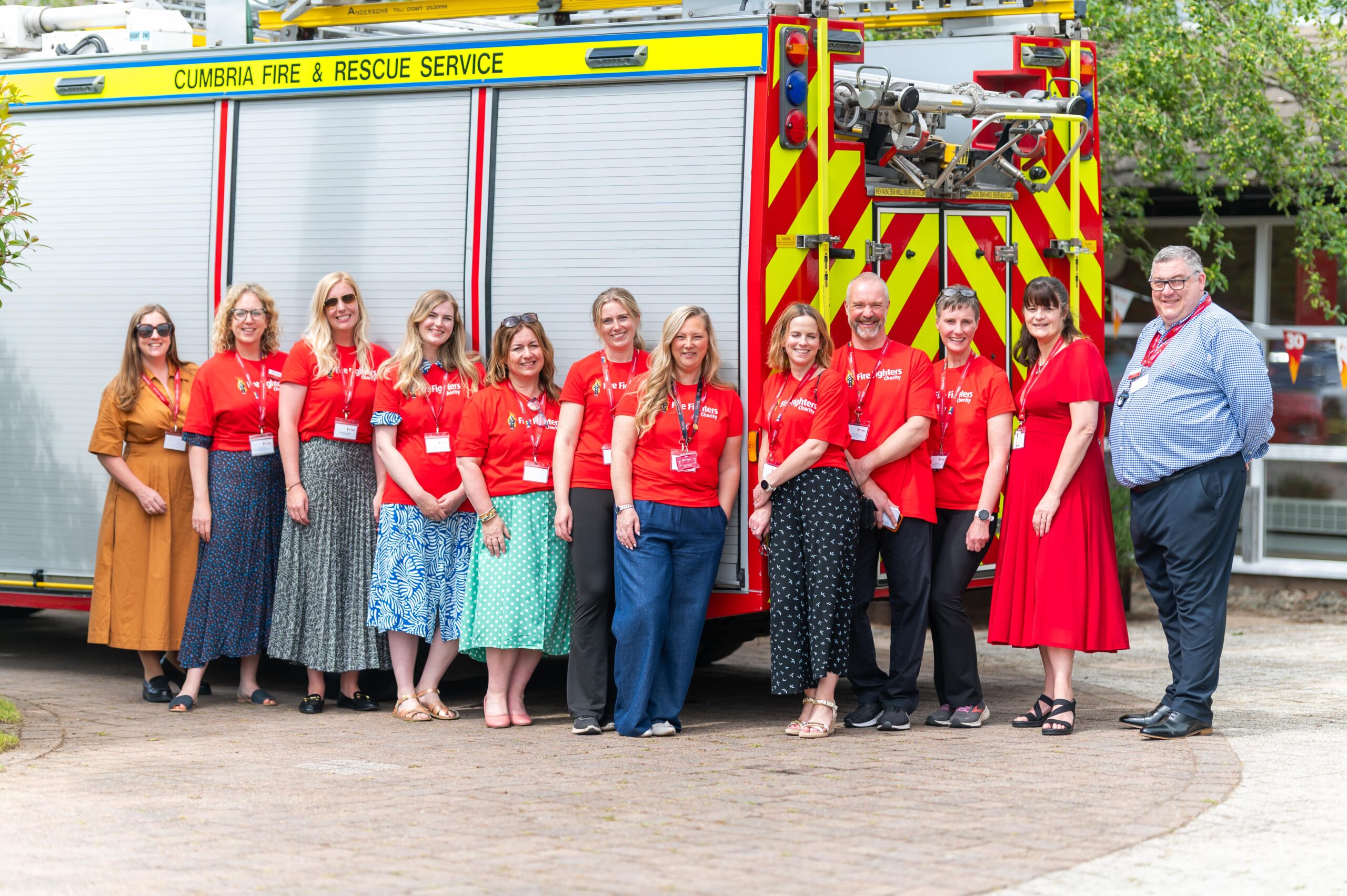 A group of twelve adults stand in front of a Cumbria Fire & Rescue Service truck. Most wear red shirts and ID badges, smiling at the camera on a sunny day. Trees and a building are in the background.