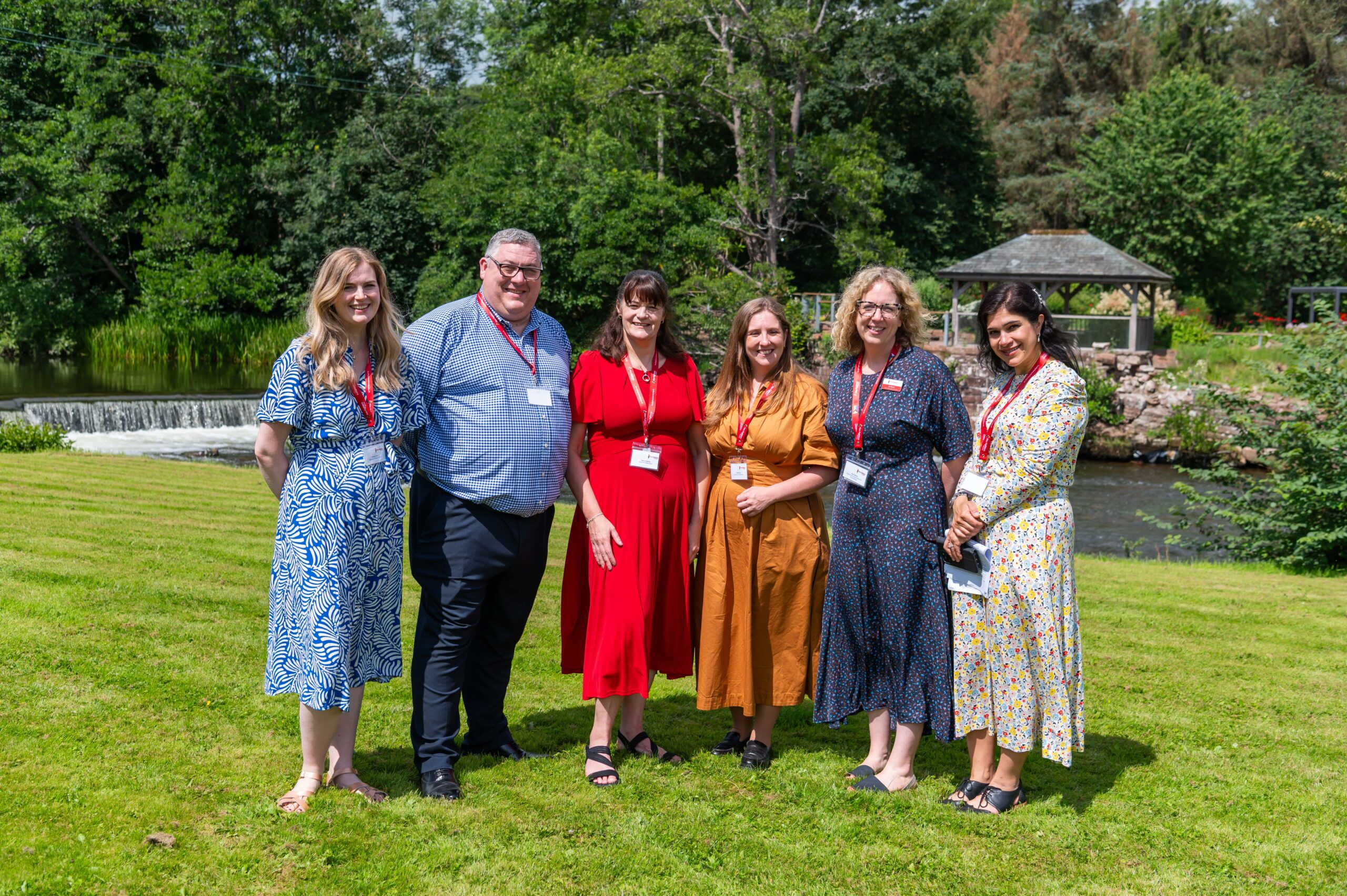 Six people stand on grass in front of a river and trees, wearing name badges and colorful outfits. They smile at the camera on a sunny day, with a small pavilion visible in the background.