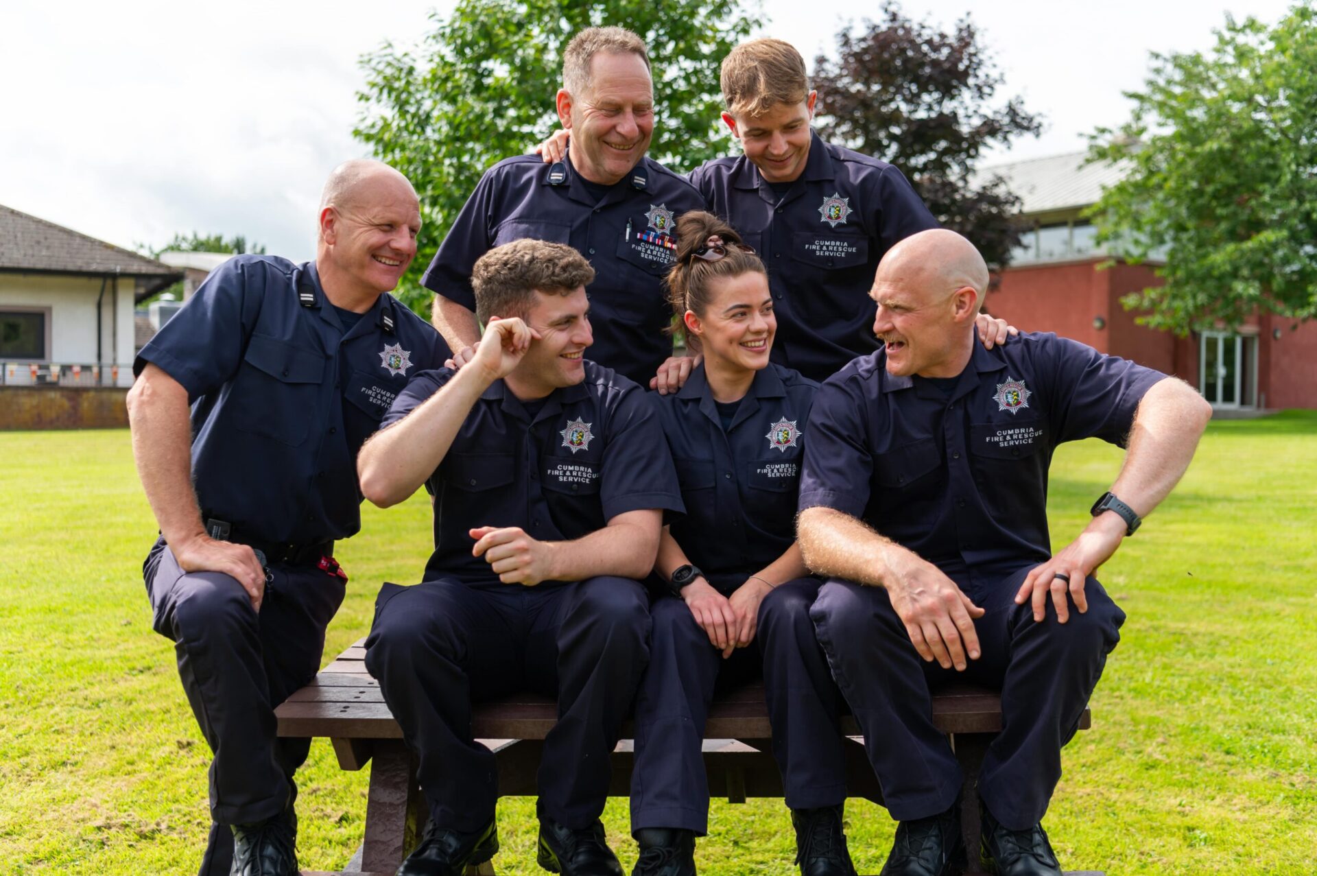 Six firefighters in uniform sit and stand around a bench outdoors on grass, smiling and laughing together, with trees and a building in the background.