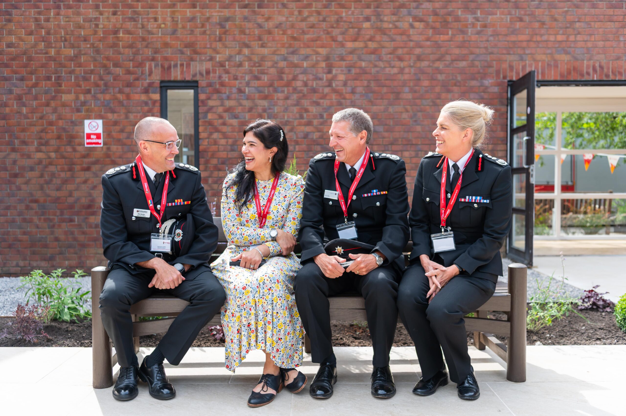 Four people sit on a bench outside a brick building, three in formal uniforms with medals and one in a floral dress. They are smiling and talking, all wearing red lanyards with name badges.