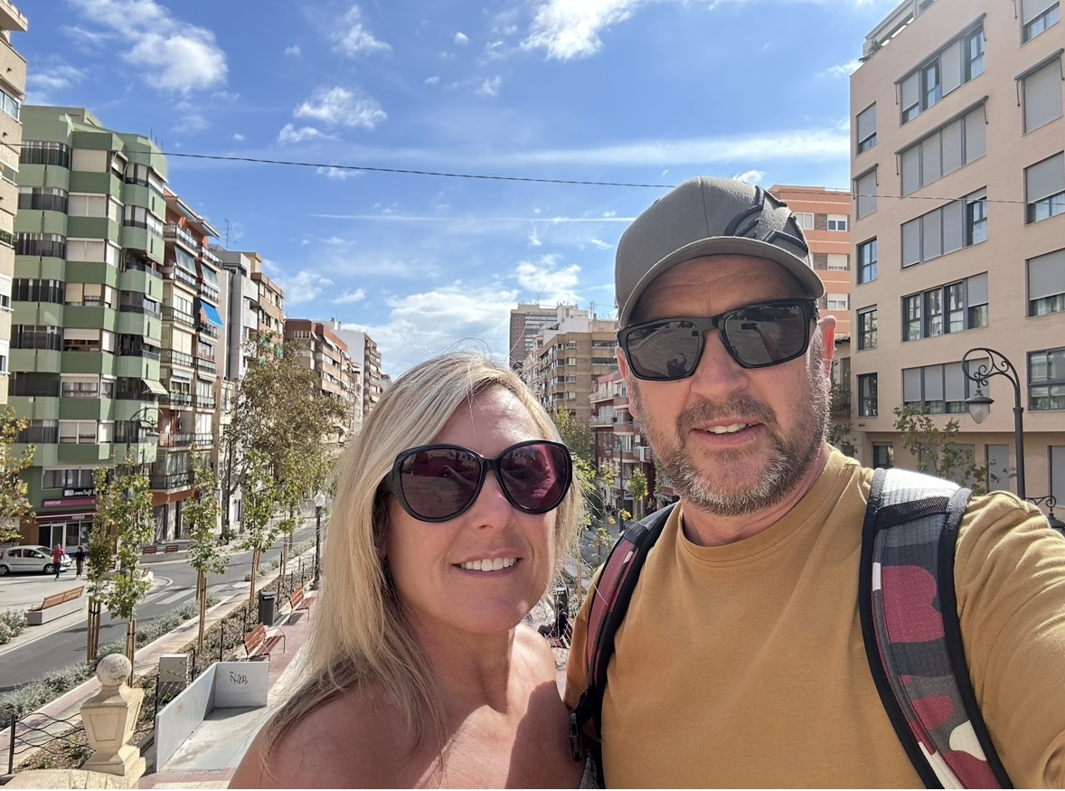 A smiling couple wearing sunglasses takes a selfie outdoors on a sunny day, surrounded by tall apartment buildings and a clear blue sky with scattered clouds in the background.