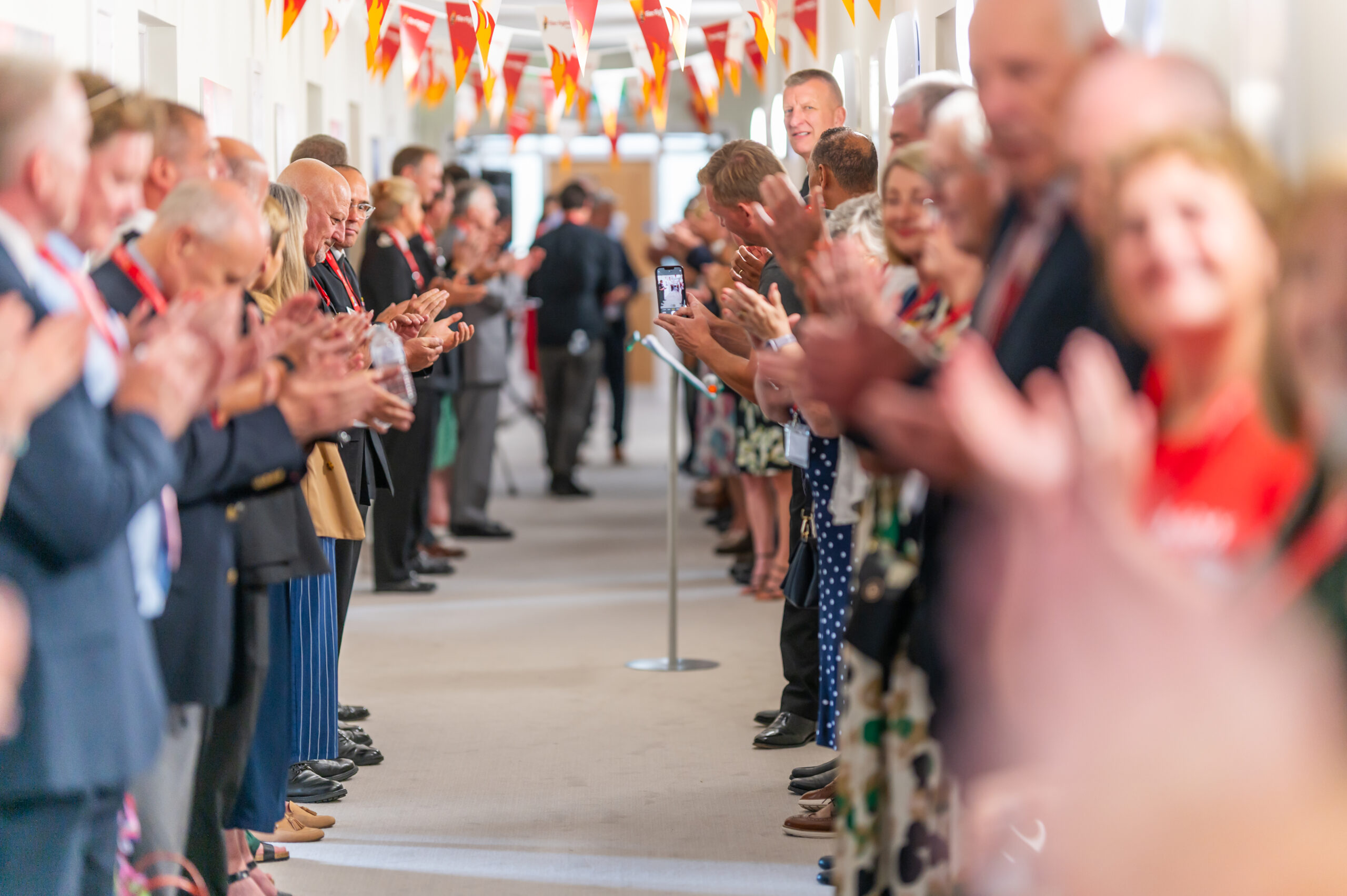 A group of people forms two lines in a hallway, clapping and smiling. Colorful triangular flags hang above them, and one person is taking a photo on a phone at the far end of the hall.