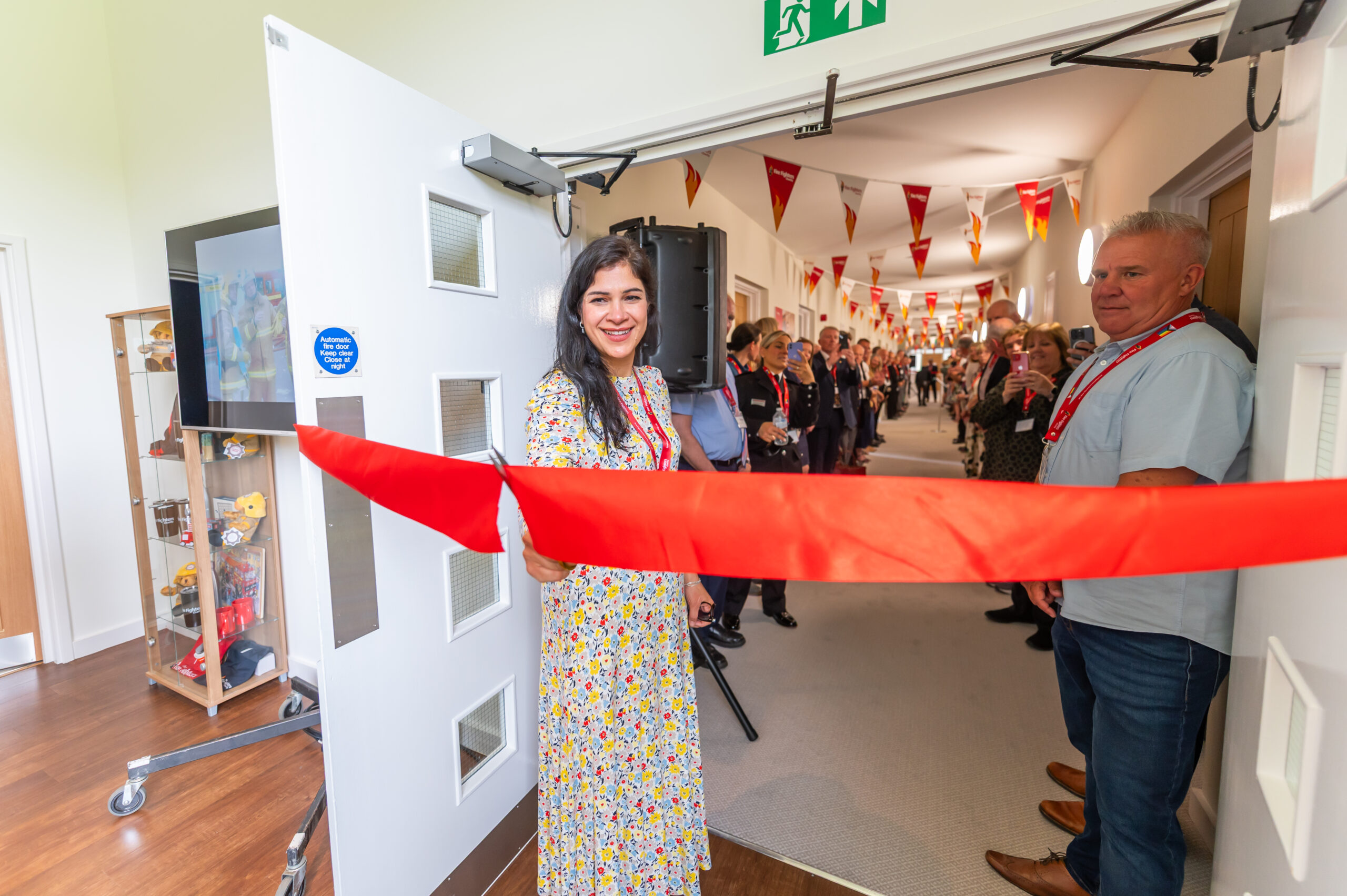 A woman in a floral dress smiles while cutting a red ribbon at the entrance of a decorated hallway, surrounded by people attending the ribbon-cutting ceremony.