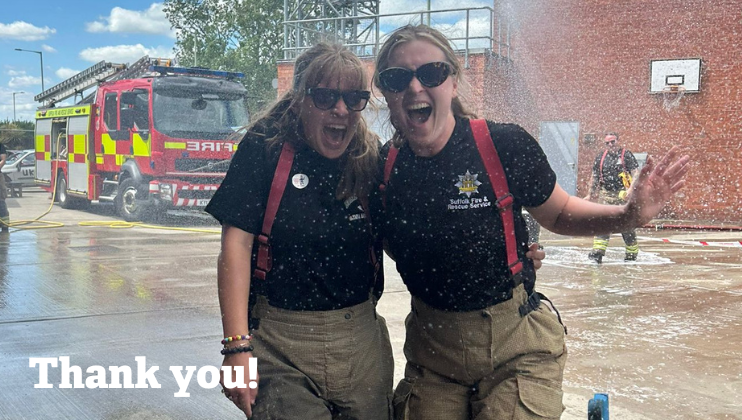 Two smiling firefighters in uniform pose together, standing in front of a fire truck and being sprayed with water. The words 
